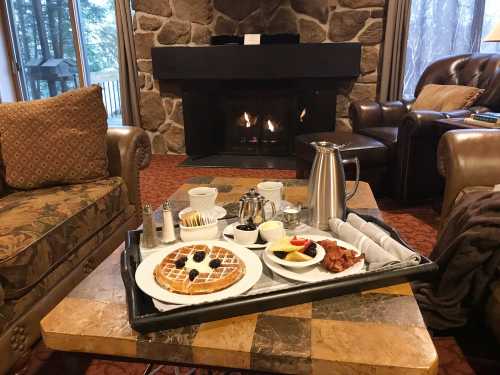 A cozy breakfast tray with waffles, fruit, and coffee in front of a stone fireplace and comfortable seating.