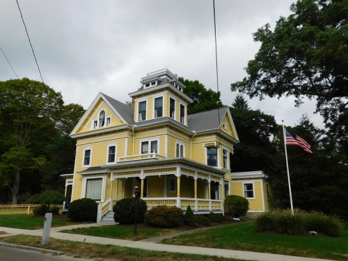 A large yellow Victorian house with a tower, surrounded by trees and a flagpole displaying the American flag.