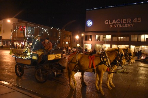 A horse-drawn carriage adorned with lights stands on a street at night, with two people inside and a distillery in the background.