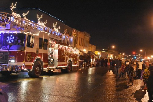 A festive fire truck adorned with lights and decorations drives past a crowd during a nighttime parade.