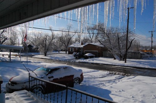 A snowy street scene with icicles hanging from a roof, a covered car, and trees blanketed in white.