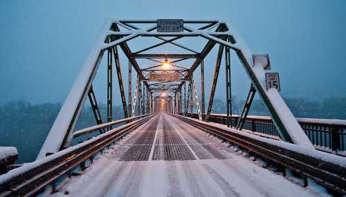 A snow-covered bridge at dusk, with streetlights illuminating the snowy path and signs visible on the structure.