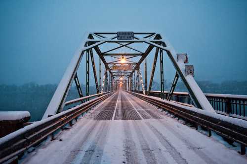 Snow-covered bridge at dusk, with streetlights illuminating the path and snowflakes gently falling.