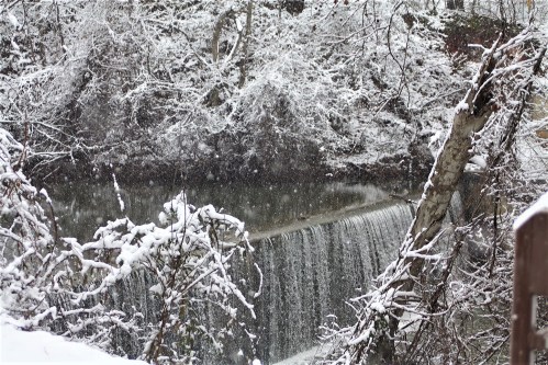 A snowy landscape featuring a waterfall surrounded by trees and a blanket of snow. Soft snowflakes are falling.