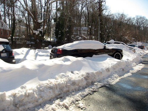 A snowy street with parked cars partially buried in snow, surrounded by trees and a clear blue sky.
