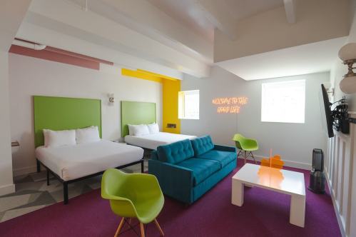 Bright hotel room featuring two beds, a teal sofa, colorful walls, and modern furniture. Neon sign reads "Welcome to the Good Life."
