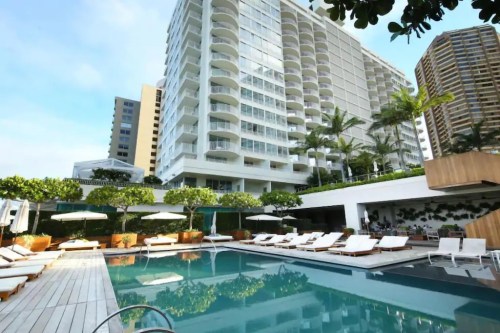 A modern hotel with a pool surrounded by lounge chairs and palm trees, set against a clear blue sky.