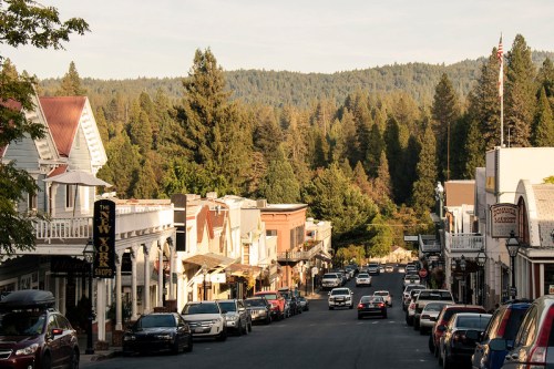A charming small town street lined with shops and parked cars, surrounded by tall trees and mountains in the background.
