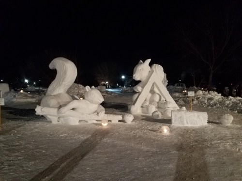 Two intricate snow sculptures at night: a squirrel and a goat, surrounded by snow and soft lighting.