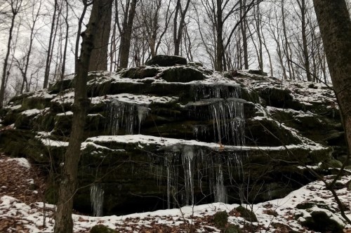 A rocky hillside covered in snow, with icicles hanging from the layers, surrounded by bare trees in a winter landscape.