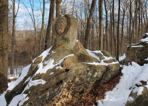 A large, weathered rock formation covered in snow, surrounded by bare trees in a winter landscape.