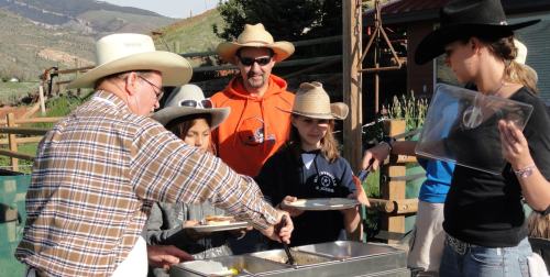 A group of people in cowboy hats serving food outdoors at a ranch, with mountains in the background.