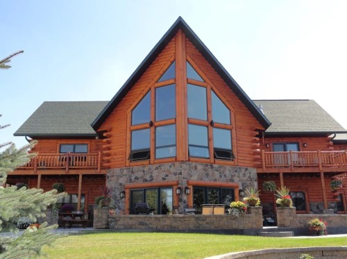 A large log cabin with a stone base, featuring large windows and a triangular roof, surrounded by greenery.