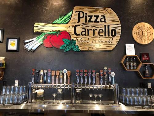 A wooden pizza peel sign with "Pizza Carrello" above a bar featuring various beer taps and glasses.