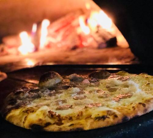 A freshly baked pizza with toppings, placed in a wood-fired oven with flames in the background.