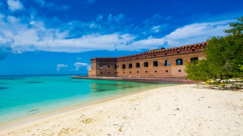 A historic brick fort on a sandy beach, surrounded by clear turquoise water and blue skies with scattered clouds.