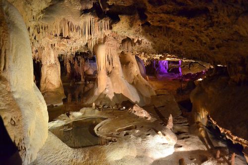 A dimly lit cave with stalactites and stalagmites, featuring a reflective pool and colorful lighting in the background.