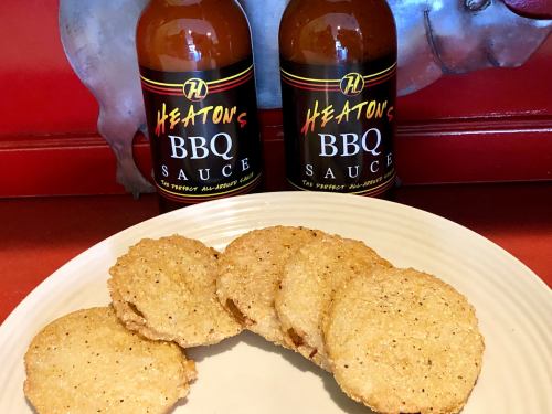 Two bottles of Heaton's BBQ sauce behind a plate of golden, crispy fried snacks.