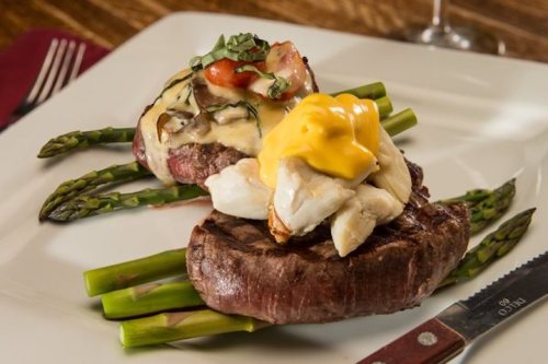 A gourmet dish featuring two grilled steaks topped with crab and cheese, served on a bed of asparagus.