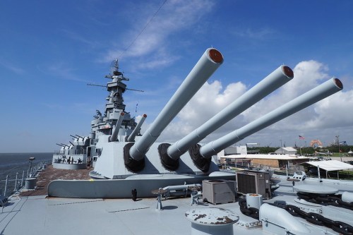 A battleship's deck featuring large cannons, with a clear blue sky and water in the background.