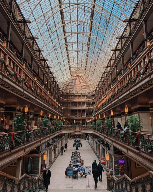 A grand indoor atrium with a glass ceiling, featuring multiple levels, greenery, and people enjoying the space.