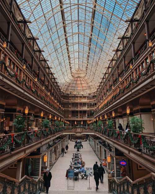 A grand indoor atrium with a glass ceiling, featuring multiple levels, greenery, and people enjoying the space.