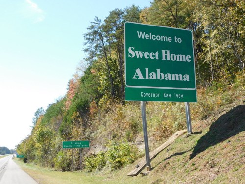 Welcome sign for Alabama, featuring "Sweet Home Alabama" and Governor Kay Ivey's name, surrounded by trees.