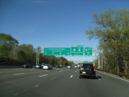Highway scene with multiple lanes, green directional signs for Garden State Parkway and Holland Tunnel, clear blue sky.