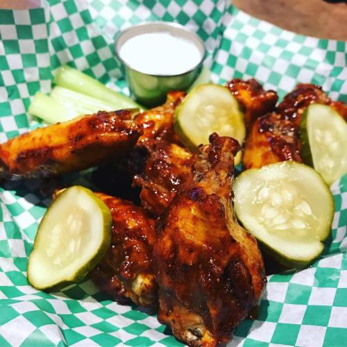 A plate of chicken wings with pickles and celery, served with a small cup of ranch dressing on a checkered paper.