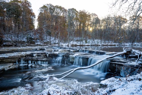 A serene winter scene featuring a waterfall surrounded by snow-covered rocks and trees at sunset.