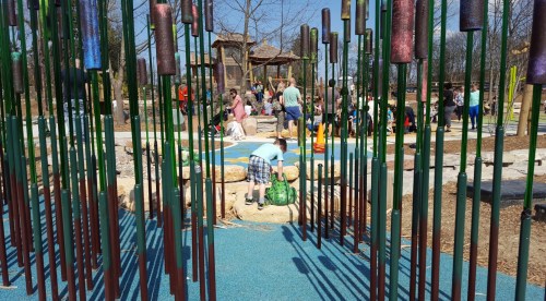 A playground scene with children playing among colorful poles and rocks, surrounded by families enjoying the outdoors.