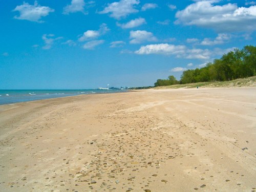 A sandy beach stretches along a calm lake under a blue sky with scattered clouds and greenery in the background.