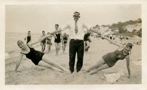 A vintage photo of two women in swimsuits balancing on the beach, holding hands with a man in a suit and tie.