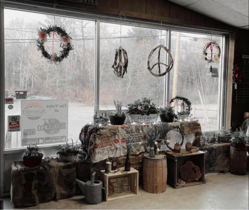 A cozy shop display featuring wreaths, plants, and rustic decor by a large window with a view of the outdoors.
