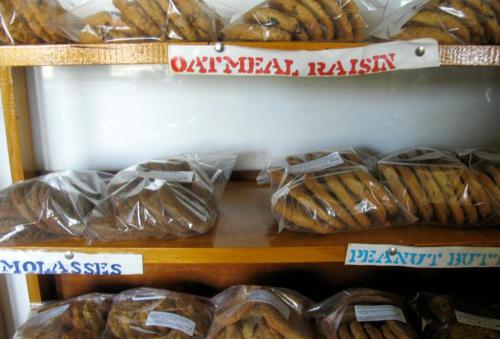 A wooden shelf displaying various packaged cookies labeled oatmeal raisin, molasses, and peanut butter.