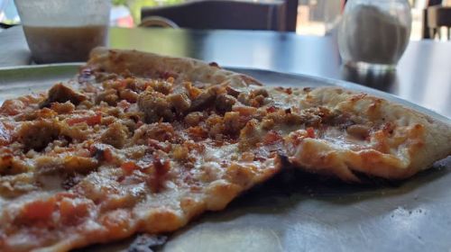 Close-up of a slice of pizza topped with meat, on a plate with a blurred background of a café setting.