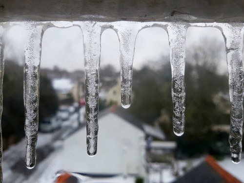 Icicles hang from a ledge, with a blurred winter landscape and houses visible in the background.