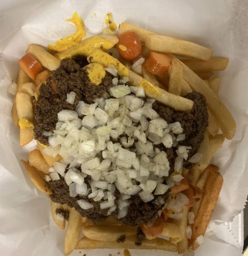 A plate of fries topped with chili, chopped onions, and mustard, served in a white paper container.
