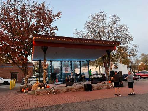 A stage set for a performance, surrounded by autumn trees and decorations, with a few people watching nearby.