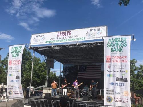 A band performs on stage at the Ambler Arts & Music Festival, with banners for sponsors and an American flag in the background.