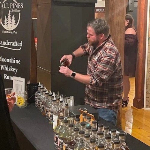 A man in a plaid shirt samples spirits at a distillery, surrounded by bottles on a bar.