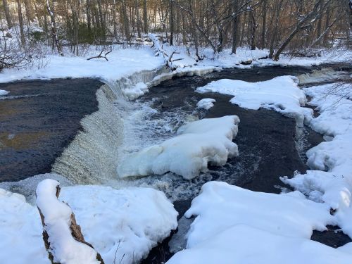 A snowy landscape featuring a partially frozen waterfall flowing into a river, surrounded by trees.
