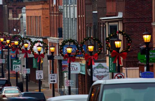 Street view decorated for the holidays, featuring wreaths and bows on lamp posts, with shops lining the road.