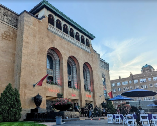 Historic building with arched windows, outdoor seating, and flags, set against a clear sky.