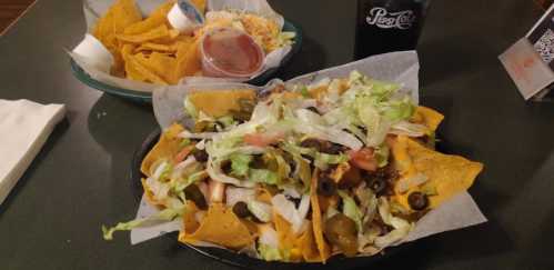 A plate of loaded nachos topped with lettuce, tomatoes, olives, and jalapeños, alongside a bowl of tortilla chips and a drink.