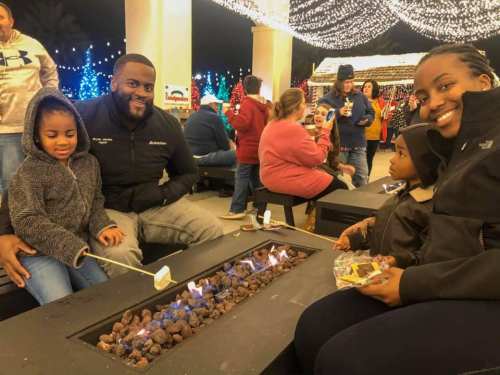 A family enjoys a festive evening by a fire pit, roasting marshmallows and surrounded by colorful holiday lights.