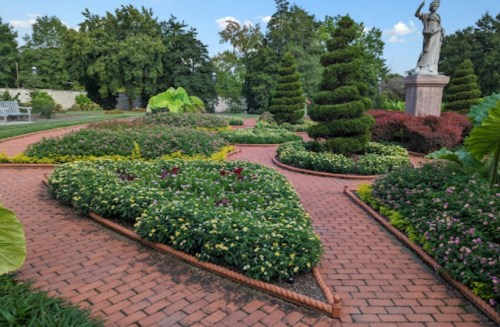 A landscaped garden with vibrant flowers, neatly trimmed bushes, and a statue in the background against a blue sky.