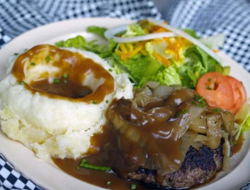 A plate featuring a hamburger topped with onions and gravy, mashed potatoes, and a side salad with tomato and cheese.