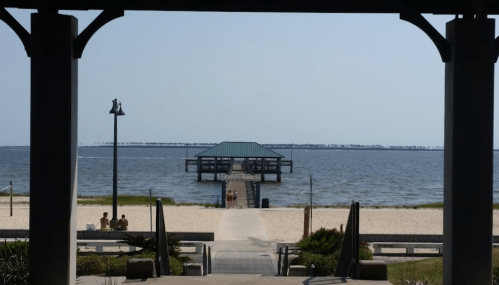 A view of a pier extending into the water, framed by a structure, with sandy beach and people sitting nearby.