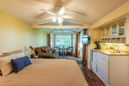 Cozy room with a bed, seating area, and kitchenette, featuring large windows and warm yellow walls.